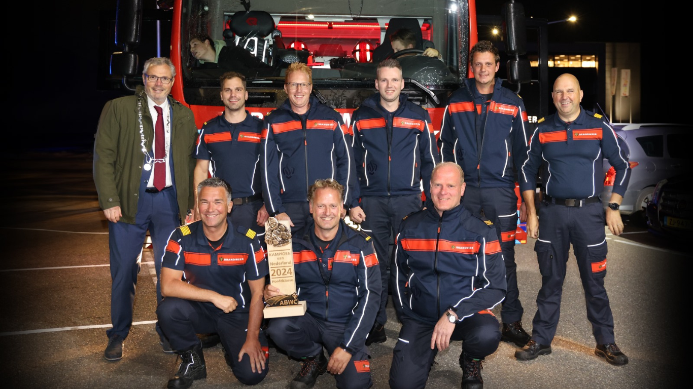 Een groep brandweermannen poseert trots voor een brandweerwagen, met de burgemeester naast zich. Ze dragen donkerblauwe uniformen en rode accenten. Twee mannen vooraan hurken, terwijl één van hen een trofee vasthoudt met de tekst 'Kampioen van Nederland 2024 Hoofdklasse ABWC'.