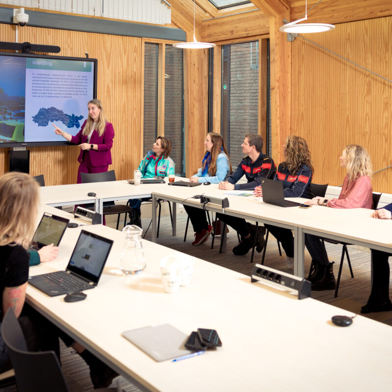 Presentatie in een vergaderruimte met houten wanden en een groot scherm. Een vrouw in een paarse blazer wijst naar het scherm, terwijl een groep deelnemers, waaronder mensen in hulpverleningsuniformen, aandachtig luistert. Enkele aanwezigen werken op laptops.